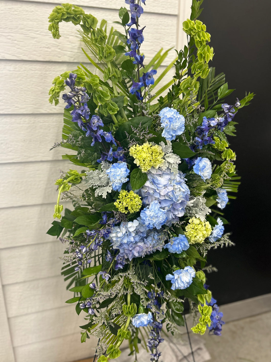 A floral easel spray arrangement featuring a mix of blue, green, and white flowers, designed for funeral services.