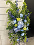 A floral easel spray arrangement featuring a mix of blue, green, and white flowers, designed for funeral services.