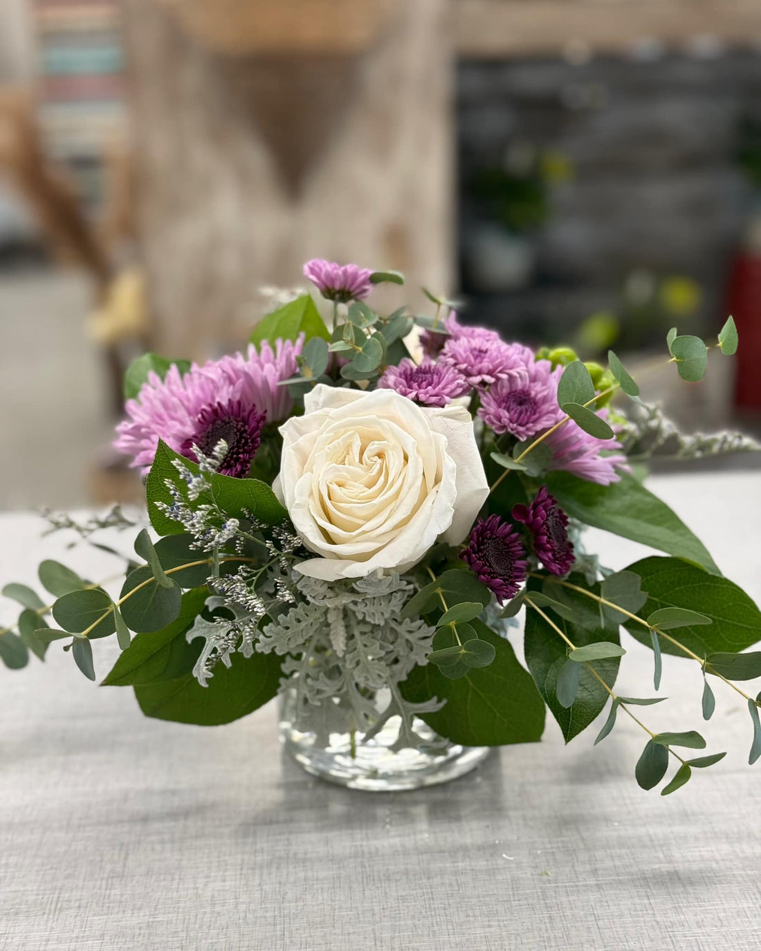 Bouquet of flowers with a white rose and purple flowers in a clear vase on a table.