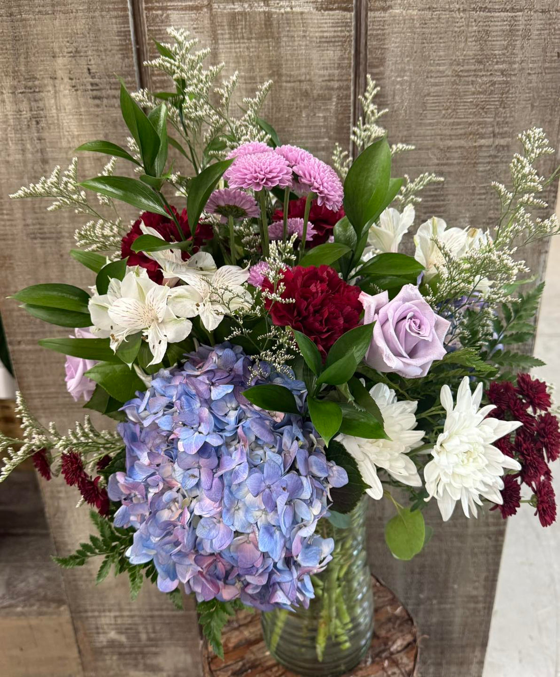 Bouquet of flowers with hydrangeas, roses, and other varieties in a clear vase against a wooden background.