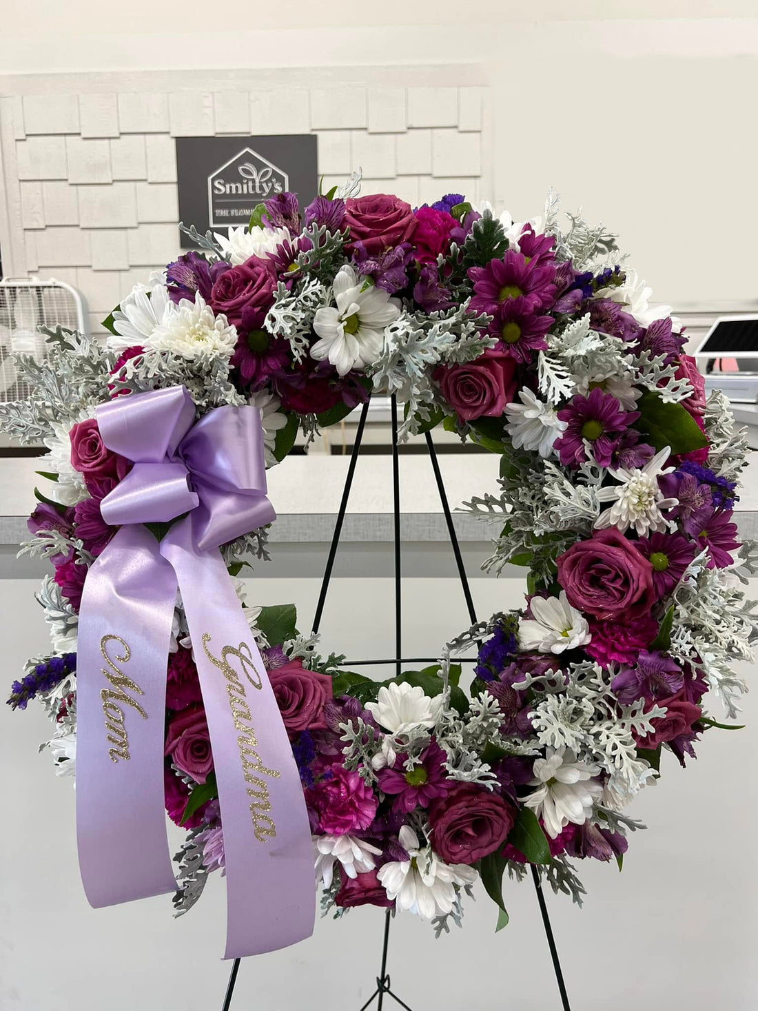 A funeral wreath made with fresh flowers, predominantly purple and white, displayed on a stand, with a purple ribbon that has the name &