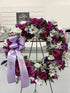 A funeral wreath made with fresh flowers, predominantly purple and white, displayed on a stand, with a purple ribbon that has the name &