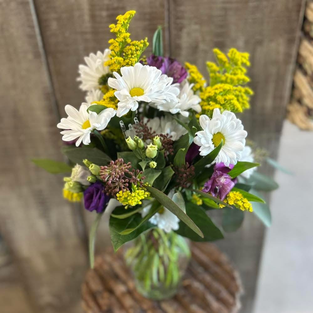 A glass vase containing a floral arrangement with white daisies, yellow flowers, and purple accents, placed on a wooden surface.