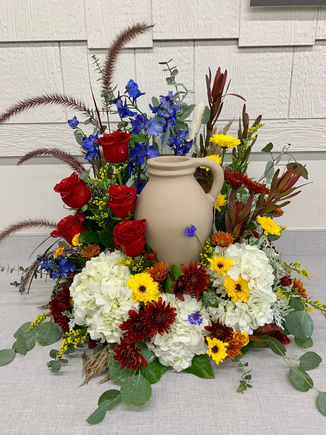 A floral arrangement featuring a ceramic urn surrounded by a variety of flowers including red roses, white lilies, and yellow daisies, with greenery and other foliage.